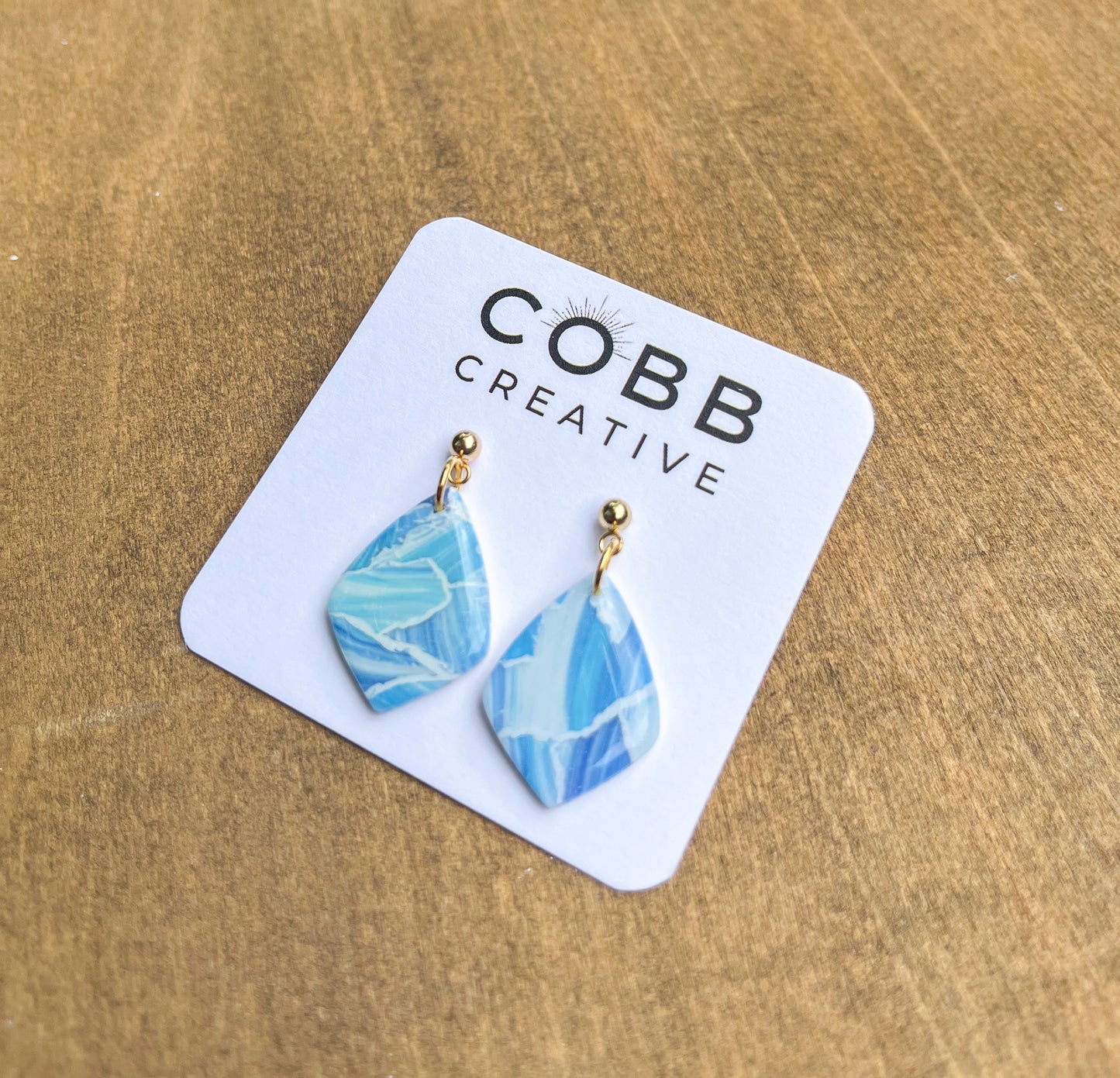 A pair of blue and white patterned polymer clay drop earrings with gold-colored ear studs, displayed on a white card with 'COBB CREATIVE' printed at the top.