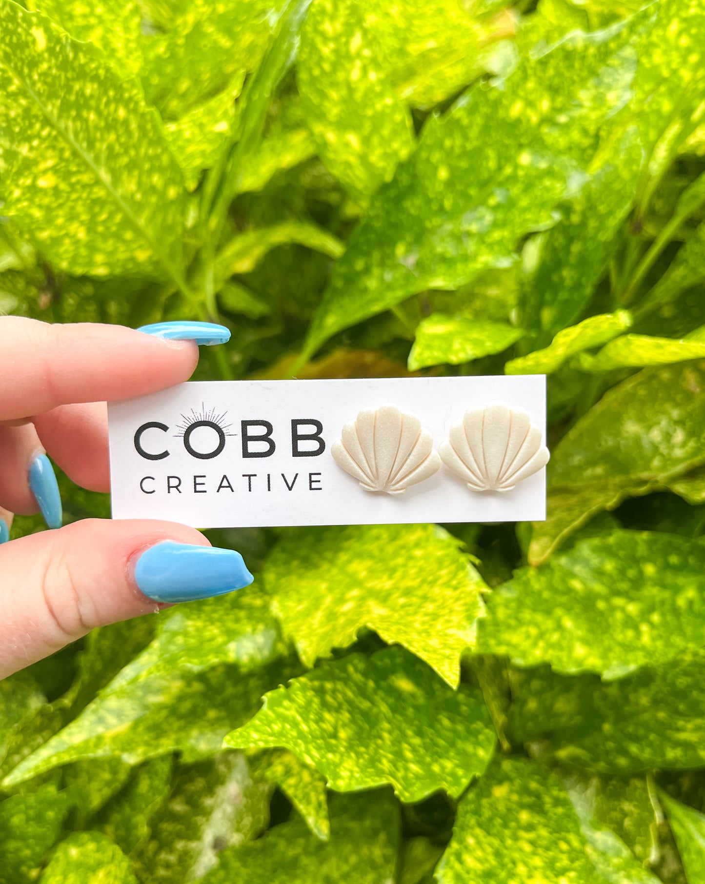 A pair of seashell-shaped stud earrings made from polymer clay, held between fingers with a green leafy background. The earrings are white and beige, and the brand 'COBB CREATIVE' is visible on a tag.