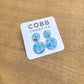 Blue marbled earrings on a Cobb Creative branded card against a wooden background