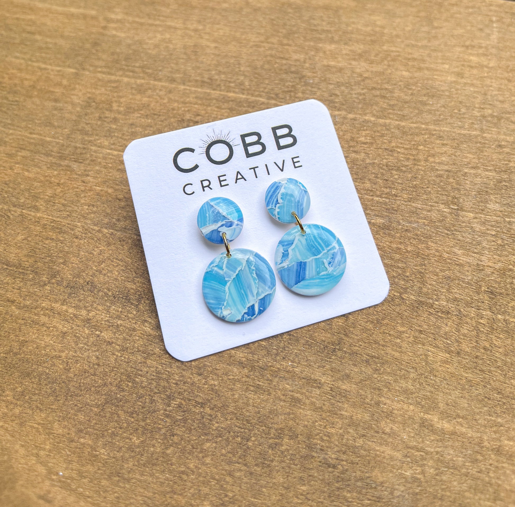Blue marbled earrings on a Cobb Creative branded card against a wooden background