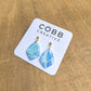 A pair of blue and white patterned polymer clay drop earrings with gold-colored ear studs, displayed on a white card with 'COBB CREATIVE' printed at the top.