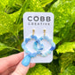 Blue earrings on a COBB CREATIVE card held against a green leafy background