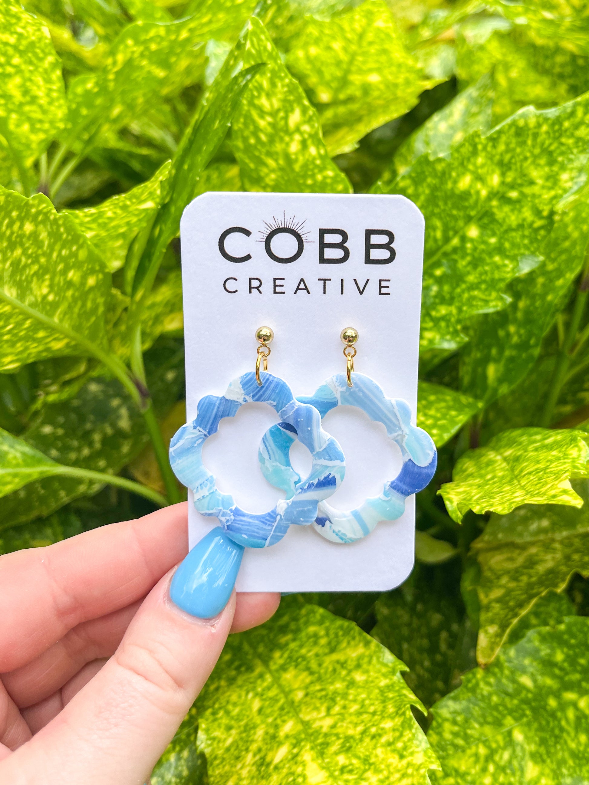 Blue earrings on a COBB CREATIVE card held against a green leafy background